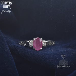 Op de afbeelding: Een zilveren ring met een ovale, dieprode edelsteen in het midden, geflankeerd door twee kleinere, ronde, heldere edelstenen. De ringband heeft een decoratief patroon. De woorden "DELIVERY DUTY paid" staan linksboven.