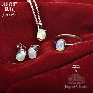 May include: A jewellery set comprising a silver necklace, ring, and earrings, each with an oval opal gemstone. The opals show blue, green, and white colours. The set is presented on a red velvet surface.