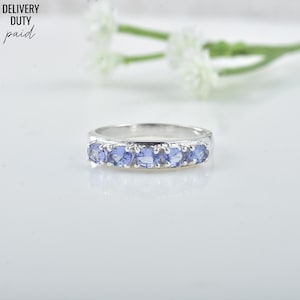 May include: A silver ring with six oval-cut, light blue gemstones set in a row. The ring is displayed on a white surface with a blurred floral background. The text "DELIVERY DUTY PAID" is in the upper left corner.