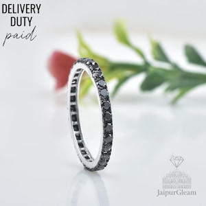 May include: A silver ring with a band of black gemstones. The ring is set against a soft white background with a blurred red flower and green leaves. Text in the upper left corner reads "DELIVERY DUTY paid".