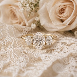 May include: A gold ring featuring a large oval-cut center diamond flanked by two smaller round-cut diamonds. The ring is set on a thin, polished gold band, showcasing a classic three-stone design with clear, faceted gemstones that reflect light.