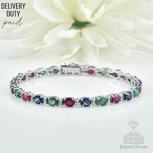 Könnte beinhalten: Ein silbernes Armband mit oval geschliffenen Edelsteinen in Rot, Blau und Grün, durchsetzt mit kleinen Diamanten. Das Armband ist vor einem weißen Hintergrund mit einer verschwommenen Blume und dem Text "DELIVERY DUTY paid" platziert.
