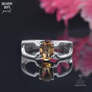 Puede incluir: Anillo de plata con una gema ovalada de color ámbar. El anillo tiene un diseño abierto único con detalles recortados en los laterales. Se puede ver el texto "DELIVERY DUTY paid". Reflejo sobre una superficie oscura.