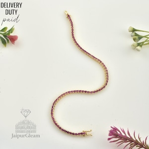 Puede incluir: Una delicada pulsera dorada con pequeñas gemas rojas, sobre una superficie blanca con detalles florales. El texto "DELIVERY DUTY paid" está en la esquina superior izquierda. El logotipo de JaipurGleam está en la esquina inferior izquierda. Una fina joya.