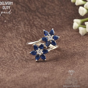 May include: A silver ring with two flower motifs, each composed of dark blue petals and a central cluster of clear stones. The ring is displayed on a brown surface. The text "DELIVERY DUTY paid" is visible in the upper left corner.