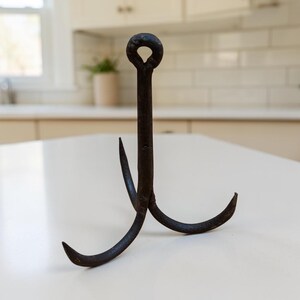 May include: A dark, weathered iron grappling hook featuring a central vertical shaft topped with a circular eyelet and three curved, pointed prongs extending outward from the base, creating a tripod-like structure with a textured, aged metal finish.