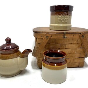 May include: A brown and white ceramic teapot, a brown and white ceramic jar, and a brown and white ceramic jar labeled "Honey Pot" are arranged on top of a brown wicker picnic basket.