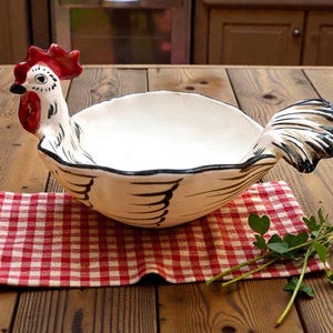 May include: A white ceramic chicken-shaped bowl with a red comb and wattle. The bowl has black detailing and is set on a red and white checkered cloth. The bowl is on a wooden table.
