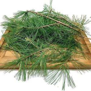 May include: A pile of fresh green pine needles on a wooden tray. The needles are long and thin, and they are arranged in a random pattern. The tray is made of light brown wood and has a smooth surface.