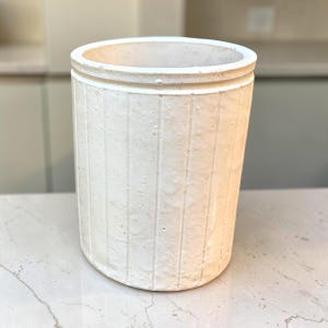 May include: A cylindrical off-white ceramic vessel featuring a textured surface with vertical ridges. The container has a thick, rounded rim and a matte finish, presenting a simple, open-top design suitable for holding various household items.