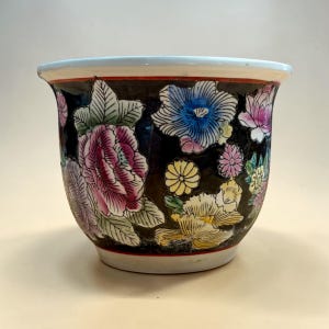 May include: A ceramic planter with a black background and colorful floral designs. The flowers are in shades of pink, blue, yellow, and purple. The rim is light blue with a red and white trim. The planter has a rounded shape.