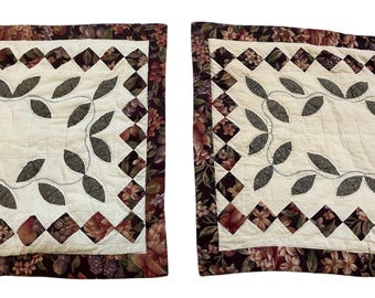 May include: Two quilted pillow covers with a floral and diamond pattern border. The center features a cream-colored background with a leaf design. The border is a dark burgundy with floral patterns.