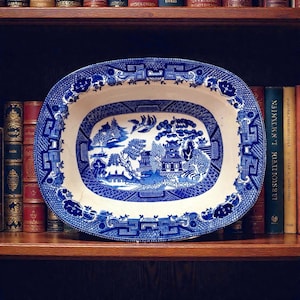 May include: Oval blue and white ceramic platter with a Willow pattern design. The platter features a detailed landscape scene in the center, surrounded by a decorative border. The dish is displayed on a wooden shelf with books in the background.
