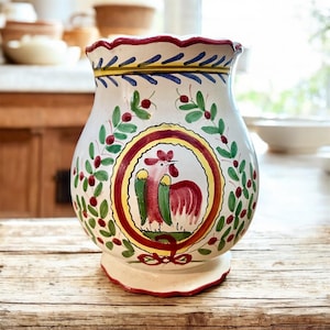 May include: A white ceramic vase with a hand-painted rooster design. The vase features a scalloped rim with red, blue, and yellow accents. Green leaves and red berries decorate the sides, and a red ribbon is at the base.