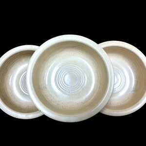 May include: Three beige-colored bowls are arranged in a row against a black background. Each bowl has a circular design in the center and a light-colored, textured appearance. The bowls are likely for serving food or decorative purposes.