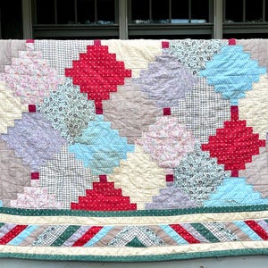 以下が含まれることがあります： パッチワークデザインの手作りキルト。赤、青、クリーム色、模様入りの布の正方形が菱形に配置されています。縁は緑色で、赤と白のストライプが入っています。暖かく、装飾的なテキスタイルです。