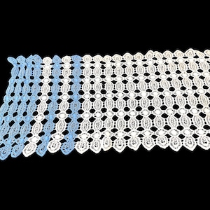 Könnte beinhalten: Weißer Tischläufer aus Spitze mit einem aufwendigen Lochmuster. Der Läufer hat einen gewellten Rand und ein sich wiederholendes ovales Muster. Die Spitze hat einen leichten blauen Farbverlauf auf der linken Seite, wodurch ein subtiler Farbkontrast entsteht.