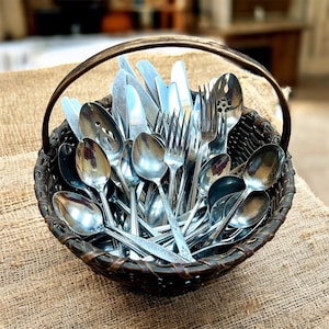 May include: A woven basket filled with an assortment of silverware. The basket contains spoons, forks, and knives, all with shiny, reflective surfaces. The basket has a handle and sits on a burlap surface.