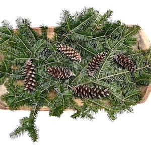 May include: A wooden bowl filled with fresh green fir branches and several brown pine cones. The arrangement is a natural, rustic decoration, perfect for the holiday season or adding a touch of nature to any space.