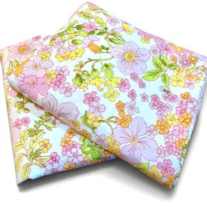 May include: A folded fabric sheet featuring a light blue background with a dense floral pattern. The print consists of various flowers in shades of pink, yellow, and orange, interspersed with green leaves and stems across the entire surface.