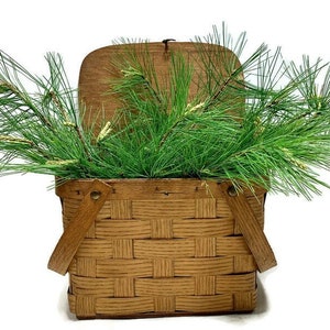 May include: A brown wicker picnic basket with a hinged lid and two handles. The basket is filled with green pine branches.