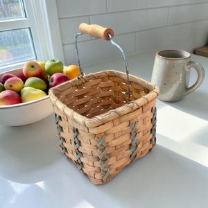 May include: A small, square woven basket with a wooden handle and gray accents. The basket is made of light brown woven material. A bowl of apples and a ceramic mug are in the background.