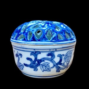 May include: A round, white and blue ceramic container with a pierced lid. The lid is a vibrant blue with decorative cutouts. The body of the container features blue floral and vine patterns. The container is set against a black background.