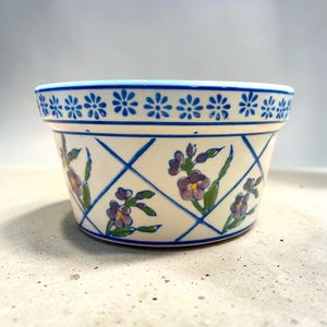 May include: A ceramic planter with a cream-colored base, adorned with a blue lattice pattern and purple floral accents. The rim features a row of small blue flower designs. A decorative piece for indoor plants.