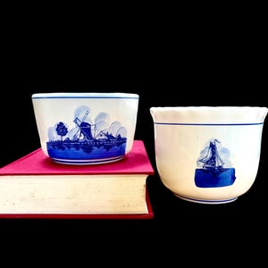 May include: Two white ceramic bowls with blue Delft-style illustrations. One bowl features a windmill scene, the other a sailboat. Both have a blue line around the rim and sit on a red book.