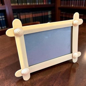 May include: A rectangular picture frame constructed from light cream-colored craft sticks. The frame features four rounded sticks arranged in a grid pattern, secured at each corner by a small, circular, spiraled decorative accent piece.