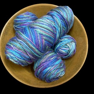 May include: A collection of colorful yarn skeins in shades of blue, purple, and teal, arranged in a golden bowl. The yarn has a variegated pattern, creating a textured appearance. Ideal for knitting and crochet projects.