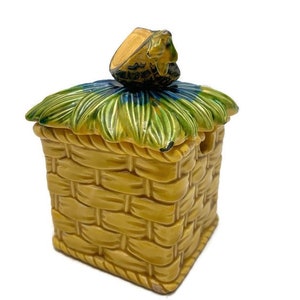 May include: A ceramic sugar bowl shaped like a thatched roof hut with a green and blue thatched roof and a small frog figurine on top.