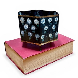 May include: A black, square container with white and blue floral designs, resting on a red book. The container has a gold trim and small feet. The book has a red cover and aged, yellowed pages.