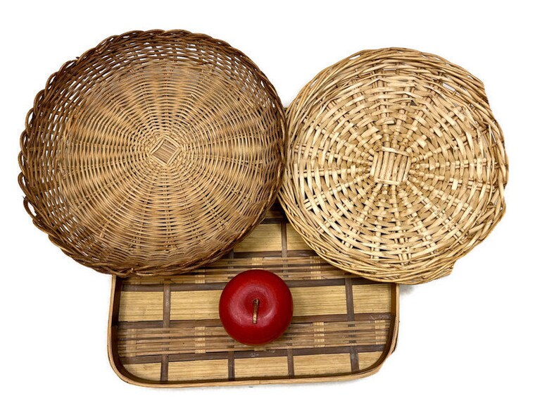 May include: Three round woven wicker baskets with a natural brown color. The baskets are arranged on a rectangular woven bamboo tray with a brown and natural color pattern. A red apple is sitting on the tray.