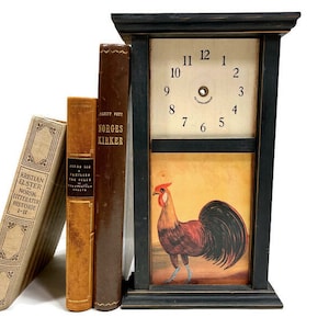 May include: A black wooden clock with a rooster illustration and a clock face with Roman numerals. Three books are stacked next to the clock, one with the title "Norges Kirker" and another with the title "Kristian Elster Norsk Litteratur Historie 1-11".