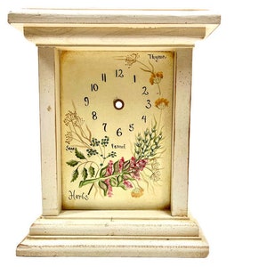May include: A white wooden clock with a cream-colored face and black Roman numerals. The clock face features a painted design of herbs and flowers, including thyme, fennel, sage, and horehound.