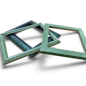 May include: Two square picture frames are layered on top of each other. The top frame is a light sage green with a textured finish. The bottom frame is a darker, muted blue-green with a decorative beaded inner edge detail and a smooth finish.