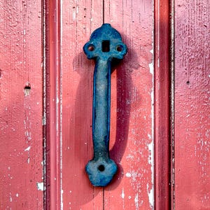 May include: A weathered, dark blue door handle with a decorative design is mounted on a red wooden door. The handle has a rectangular center and rounded ends with holes. The door's paint is chipped, revealing the wood underneath.