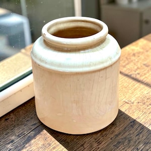 May include: A cylindrical ceramic jar featuring a glossy off-white glaze with a visible network of fine crackle patterns across the entire surface. The vessel has a wide, open mouth with a thick, rounded rim and a smooth, flat base.