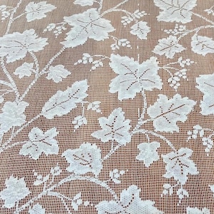May include: Close-up of a white lace fabric with a repeating pattern of grape leaves and vines. The delicate lace is set against a brown background, creating a contrast that highlights the intricate design. The fabric is likely used for home decor or crafting.