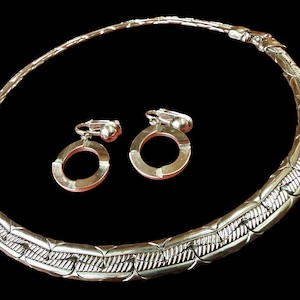 Könnte beinhalten: Silberfarbenes Halsketten- und Ohrring-Set. Die Halskette ist im Choker-Stil mit einem strukturierten, gewebten Design. Die Ohrringe sind im Clip-On-Stil und haben ein kreisförmiges Design. Das Schmuckset wird vor schwarzem Hintergrund präsentiert.
