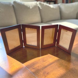 May include: A four-panel folding picture frame with a dark brown wooden finish. The frame features four rectangular openings for photographs, connected by metal hinges that allow the unit to stand upright in a zigzag or accordion configuration.