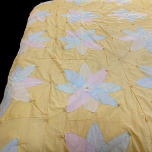 May include: A yellow quilt with large, appliqued flower designs in pastel pink, blue, and white. The flowers are evenly spaced across the quilt's surface, and the quilt is stitched with small, round, gold-colored accents.