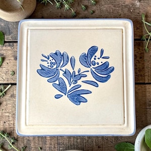 May include: Square, off-white ceramic serving tray with a blue floral design. The tray has a light blue border and a raised edge. The floral design features two large flowers and several leaves. The tray is on a wooden surface.