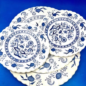May include: A collection of white ceramic plates with a blue floral design. The plates have a scalloped edge and feature a detailed pattern of flowers, leaves, and other botanical elements. The plates are arranged on a blue surface.