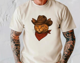 May include: Beige t-shirt featuring a cartoon cat wearing a brown cowboy hat with a star and a red bandana. The cat has orange fur and a serious expression. The shirt has short sleeves.