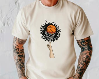 May include: Beige t-shirt featuring a basketball graphic. The design shows a basketball balanced on a finger, with a basketball net and a circle of black birds around it. The shirt has short sleeves and a crew neck.