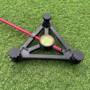 Puede incluir: Una herramienta de alineación de golf triangular negra con tres pies ajustables y un nivel de burbuja. Un palo de golf rosa brillante se inserta a través del centro de la herramienta, descansando sobre una superficie de césped verde.