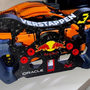 May include: A Formula 1 style steering wheel replica featuring a black carbon fiber texture, dark blue side grips, and red circular buttons. The center displays the Red Bull logo, with the text VERSTAPPEN, the number 3, and ORACLE printed on the frame.