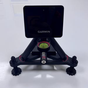 Puede incluir: Dispositivo Garmin negro montado en un trípode negro con detalles rojos. El dispositivo tiene una pantalla y el logotipo de Garmin. Un nivel y un puntero láser son visibles. El trípode tiene patas ajustables.
